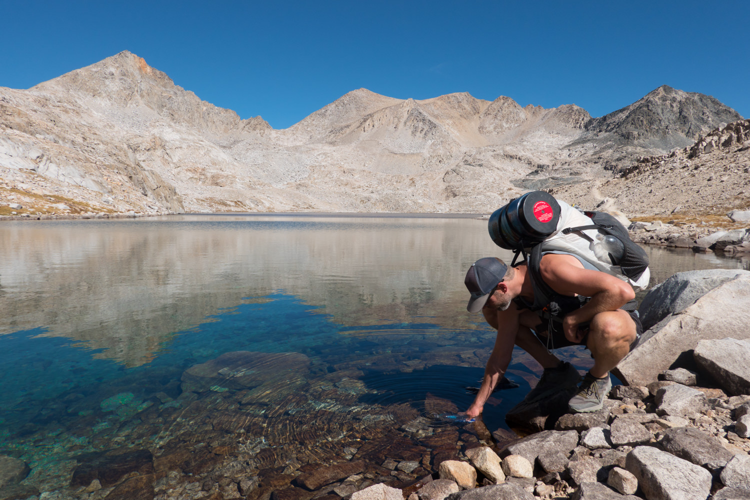 backpacker on the john muir trail bending down to fill his water bottle from a clear still lake reflecting mountains