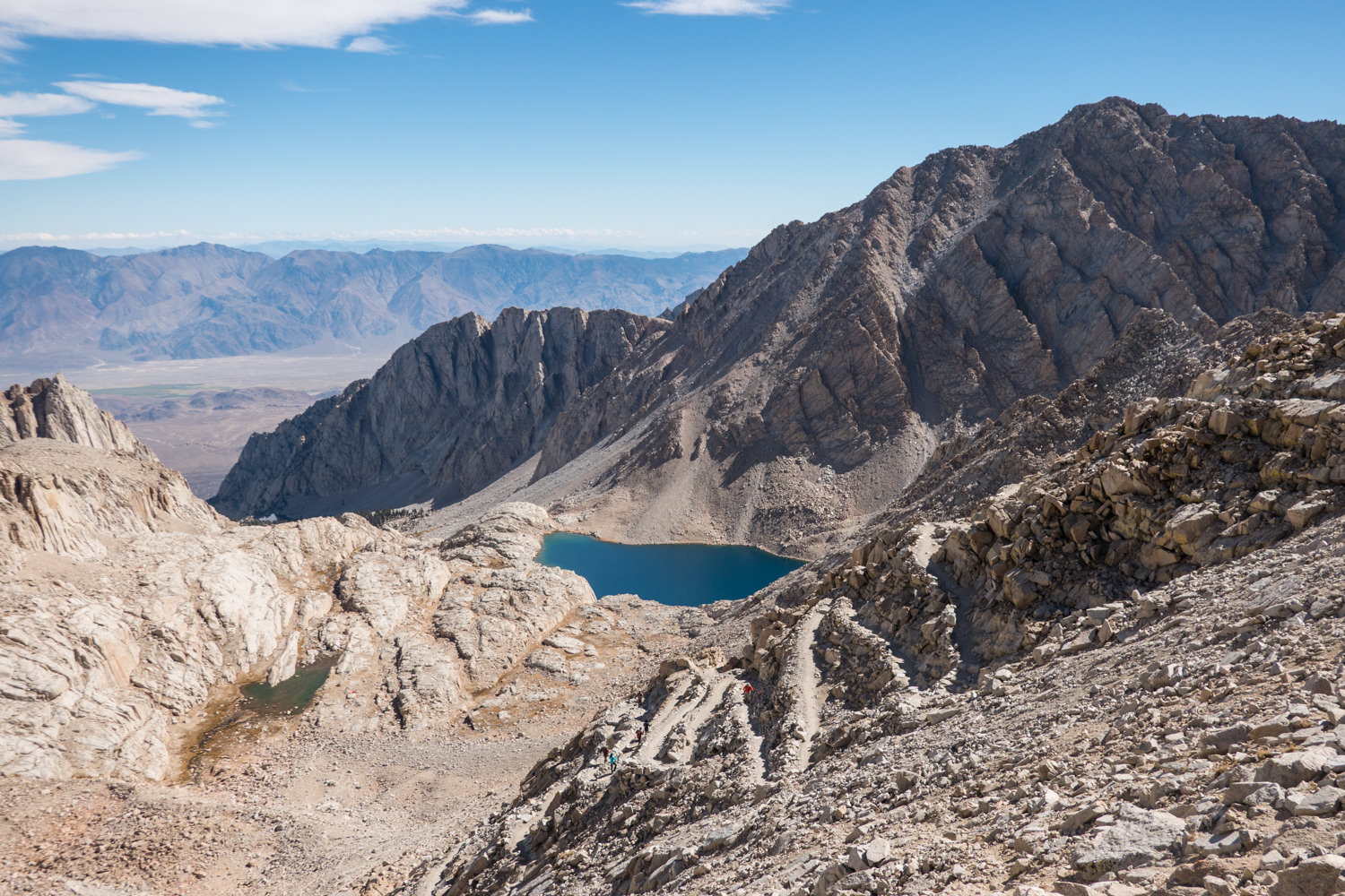 A blue tarn surrounded by mountains with the valley floor in the background. A well made trail with several switchbacks and a backpacker hiking down them in the the foreground.