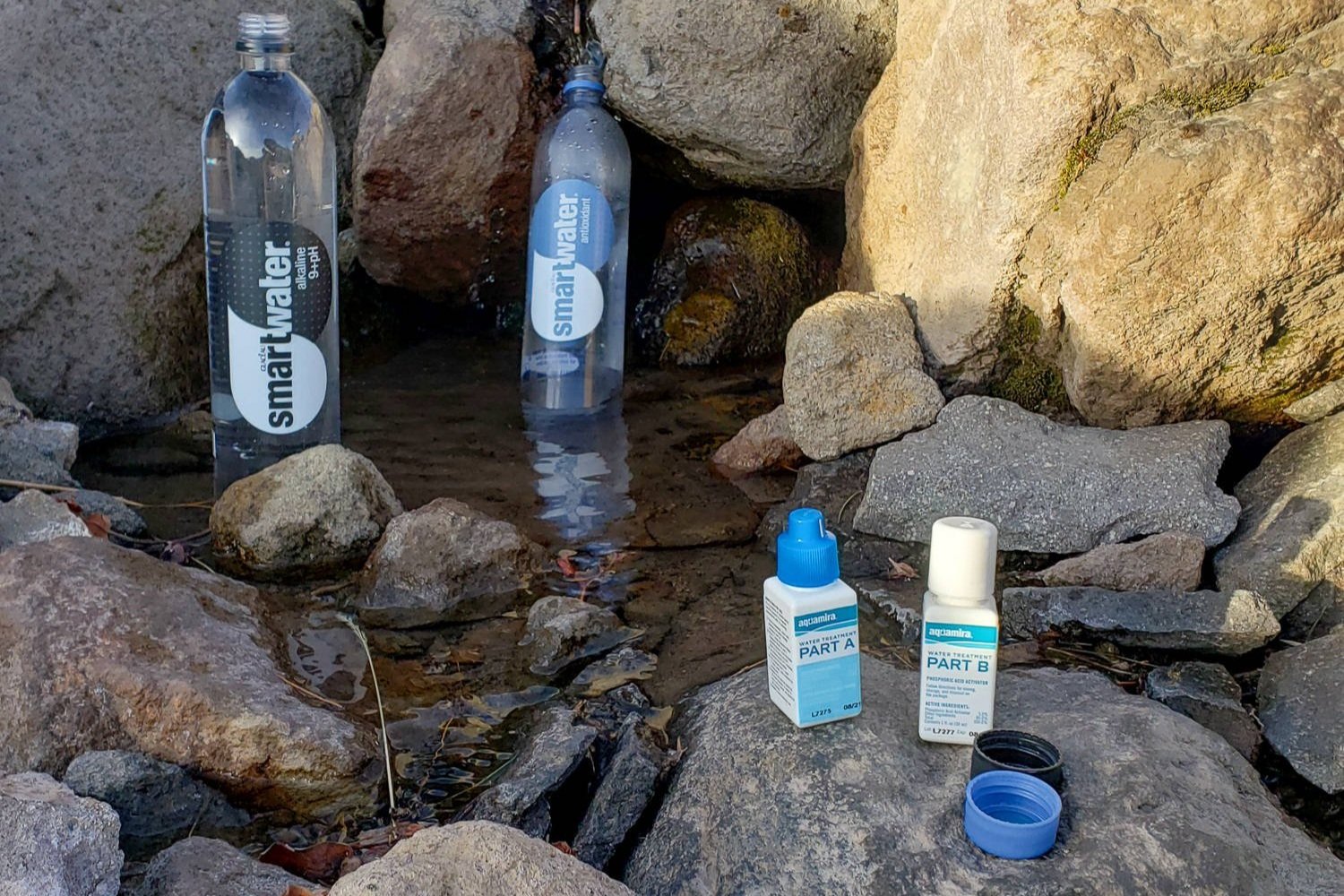Aquamira bottles sitting on a rock in front of a spring with two water bottles in it