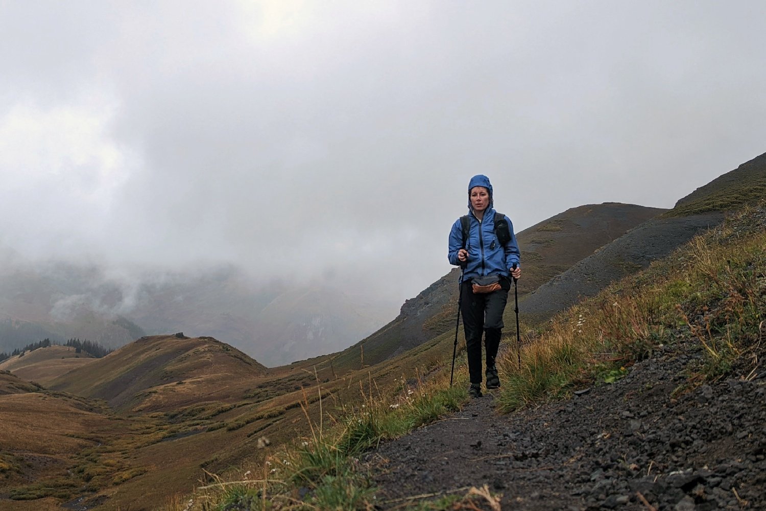 A hiker walking on a mountain trail wearing the Enlightened Equipment Visp rain jacket in the rain