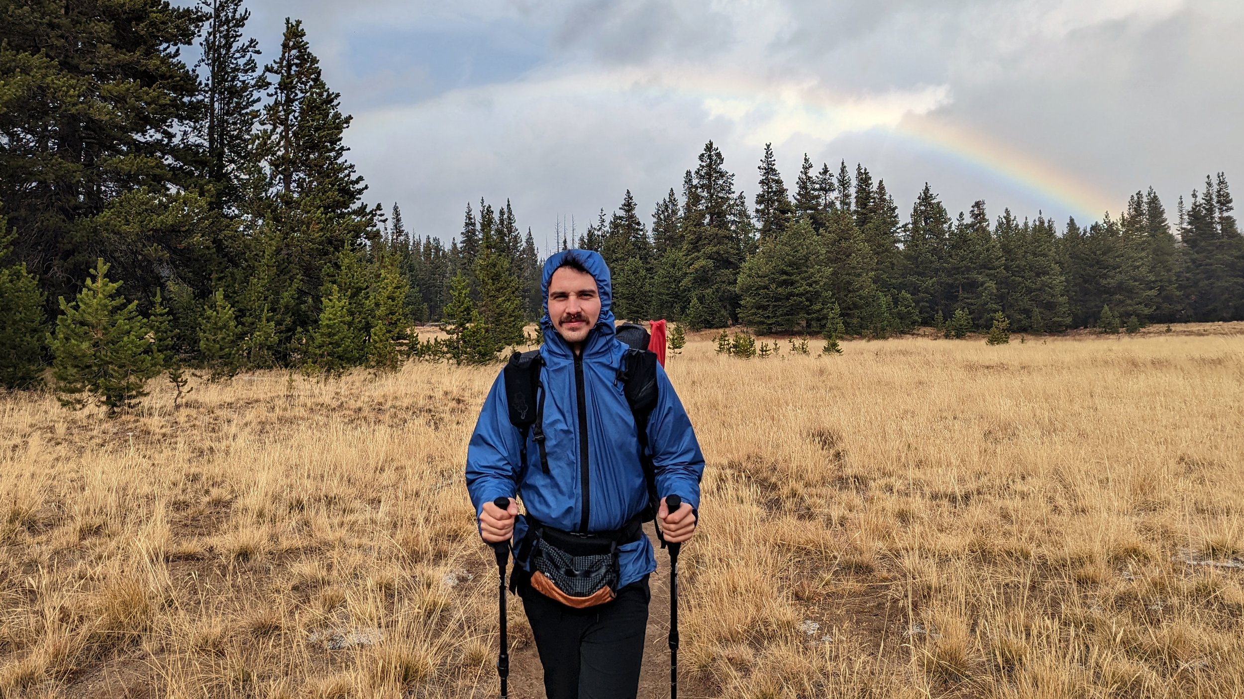 A hiker wearing the Enlightened Equipment Visp Rain jacket on a trail with a rainbow in the background