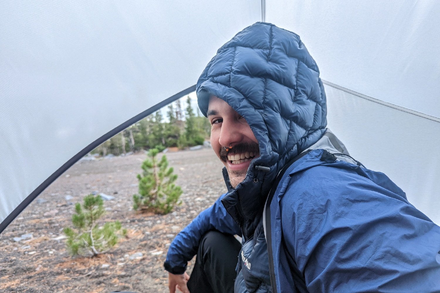 A hiker in a tent wearing the Enlightened Equipment Visp rain jacket over a puffy coat
