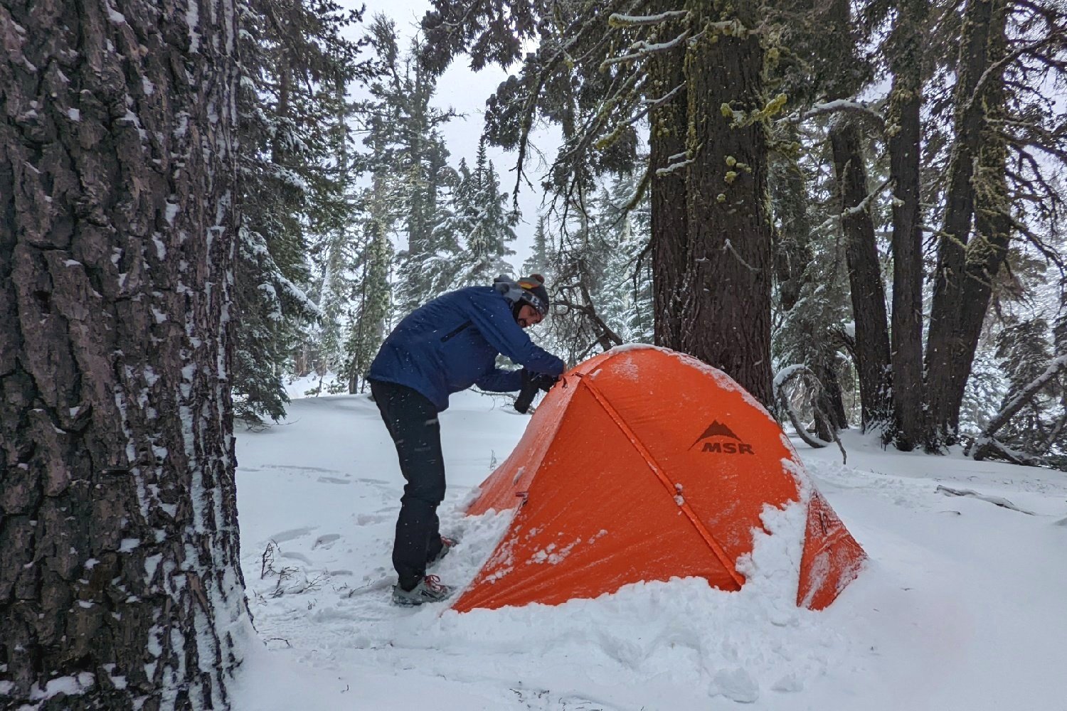 A hiker wear an Enlightened Equipment Visp rain jacket adjusting the rainfly on a tent in the snow
