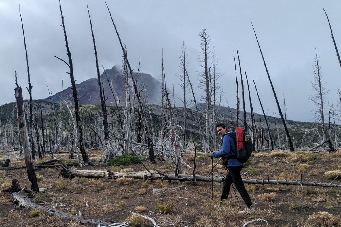 A hiker wearing the Enlightened Equipment Visp walking on a burned section of trail in front of a mountain