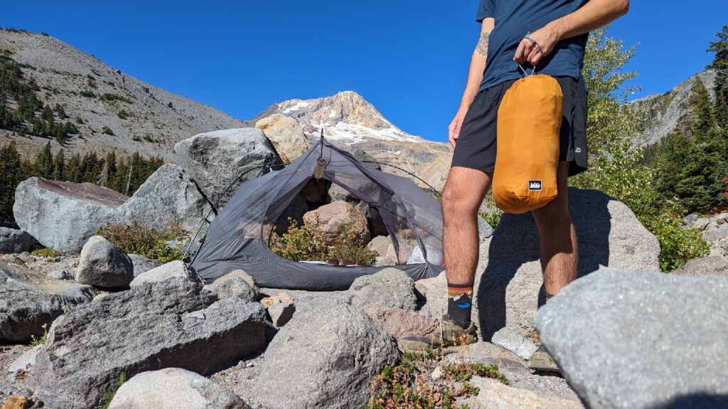 A hiker holding an orange REI Lightweight Drawstring Stuff Sack with a backpacking tent and a mountain peak in the background