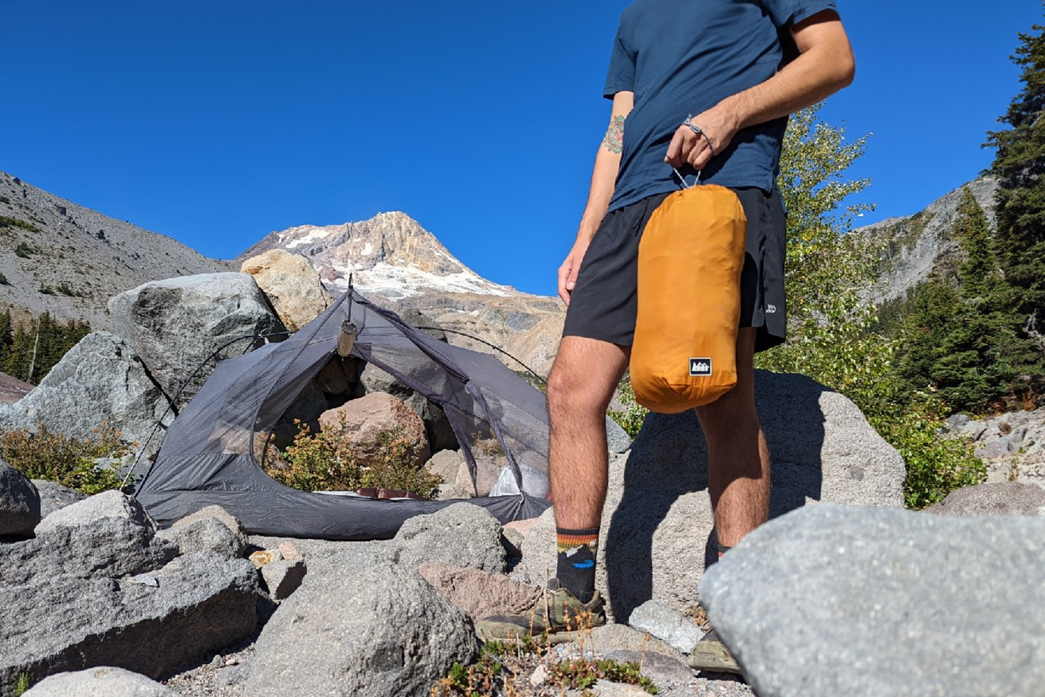 A hiker holding an orange REI Lightweight Drawstring Stuff Sack with a backpacking tent and a mountain peak in the background