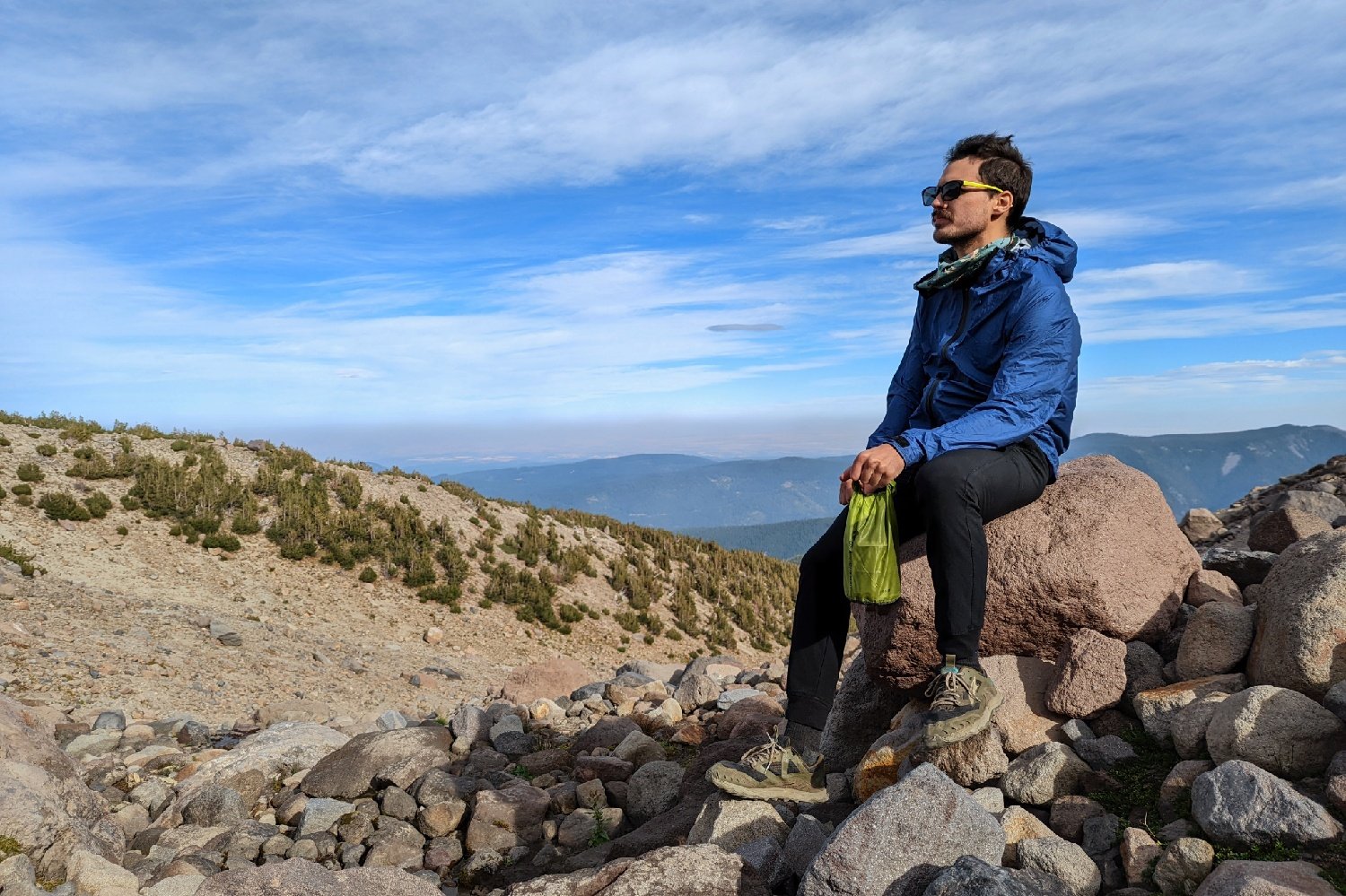 A hiker sitting on a rock in front of a mountain view wearing the Enlightened Equipment Visp rain jacket