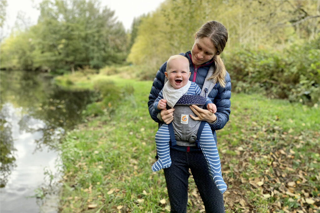A mom looking down at her smiling baby in a front carrier while hiking near a pond