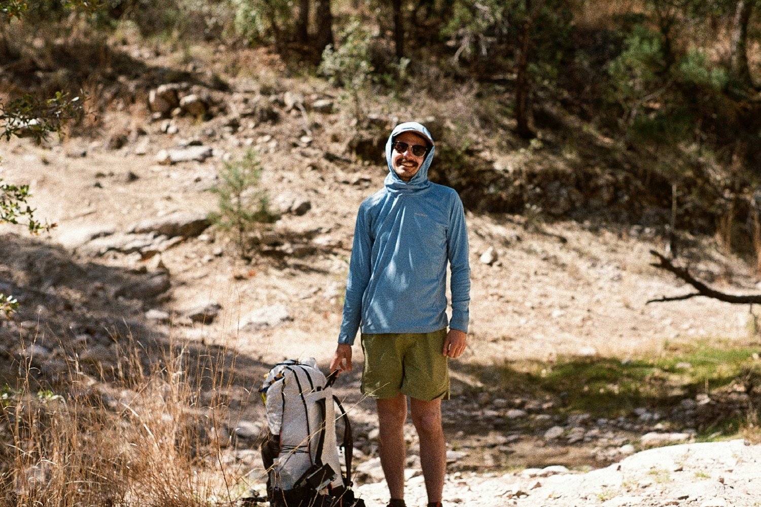 A smiling hiker standing next to their Hyperlite Mountain Gear Unbound 40 backpack with some trees and desert shrubs nearby