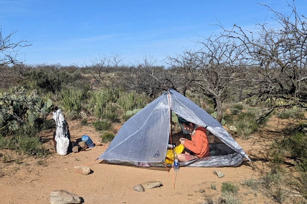 A hiker sitting inside the HMG Unbound 2 holding up a sleeping pad - the tent is in a desert campsite with one vestibule door open tent