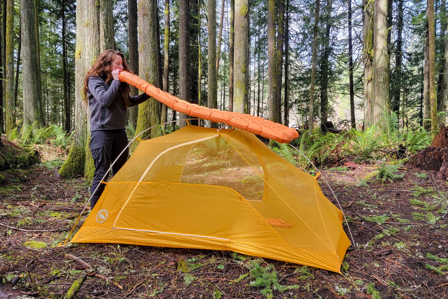 a backpacker in a forest sets up their sleeping pad while standing next to their pitched tent.