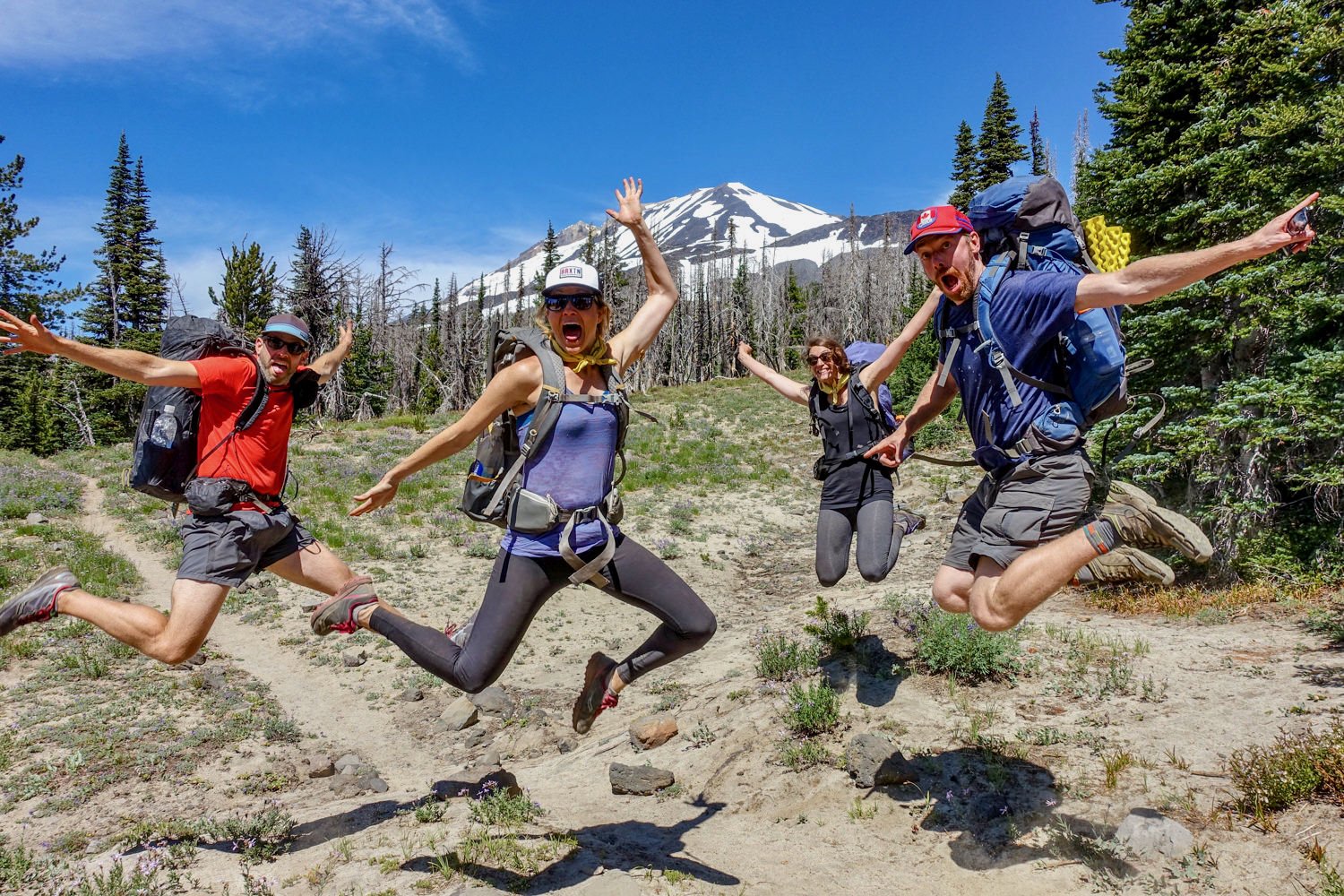 Four hikers having fun jumping high in the air above a trail with a mountain in the background