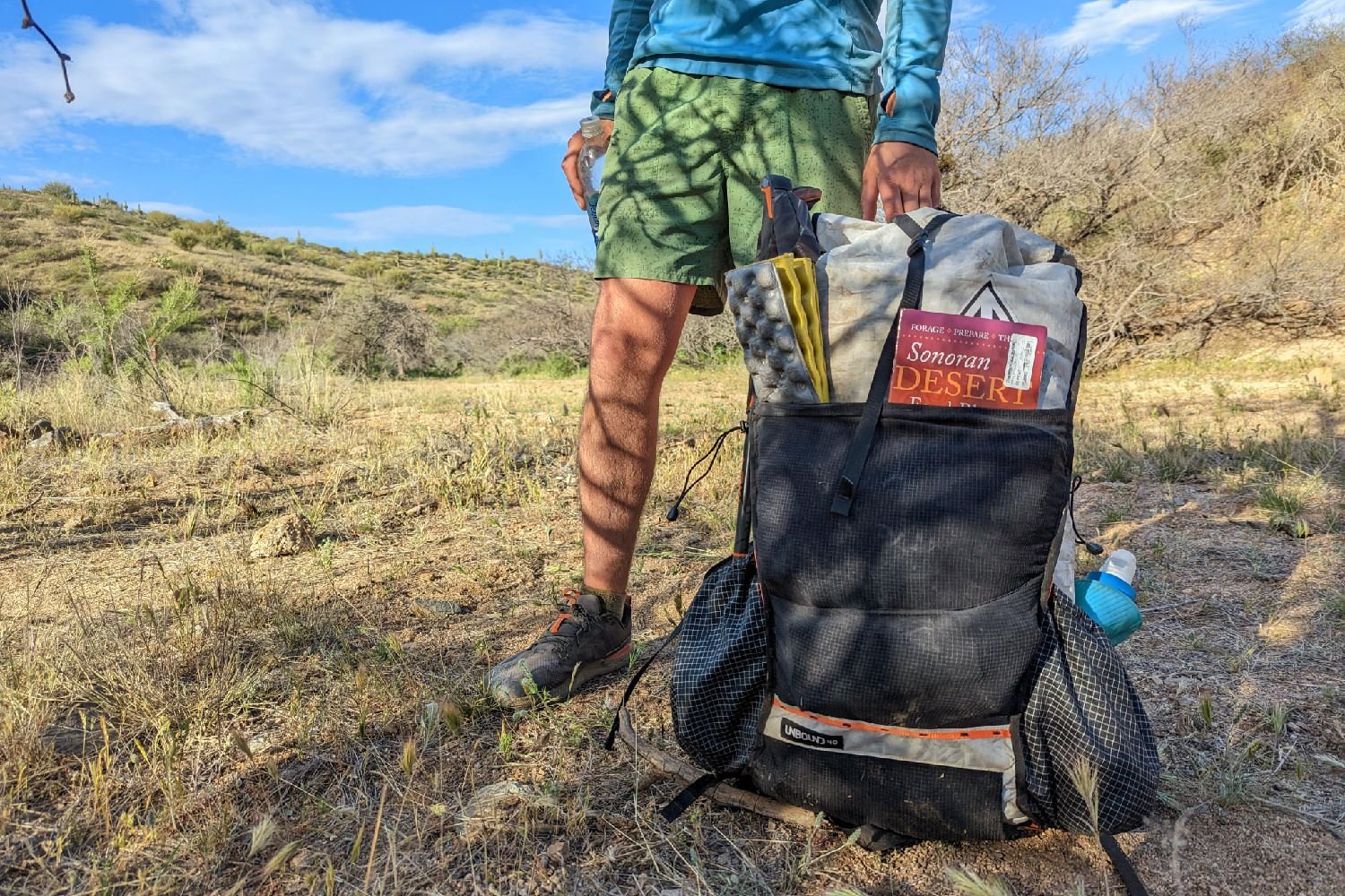 A packed Hyperlite Mountain Gear Unbound 40 sitting on the ground next to a standing hiker in a desert scene - there is a Sonoran Desert field guide and a Z seat in the front pocket