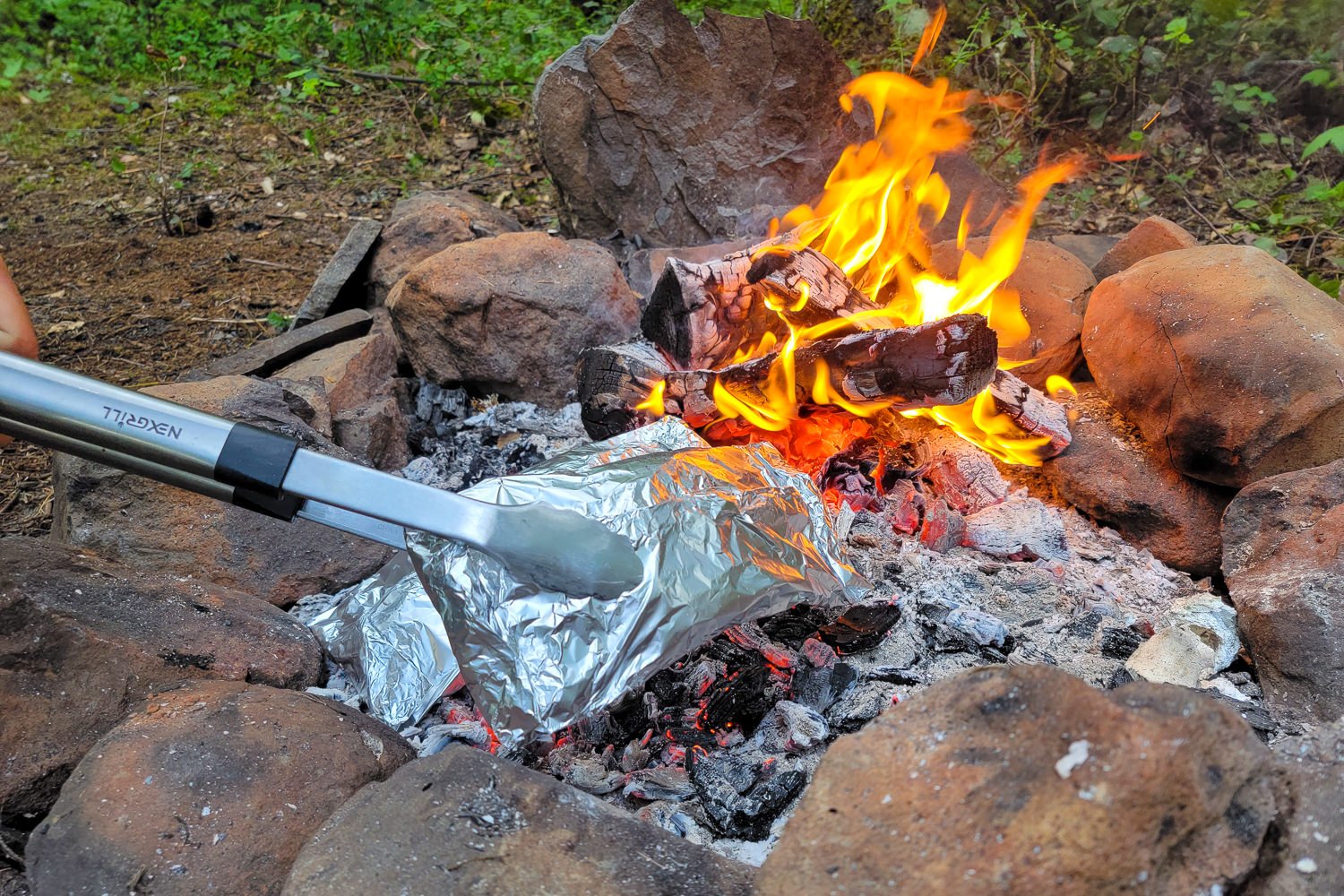 Closeup of BBQ tongs turning a foil packet meal over in a campfire