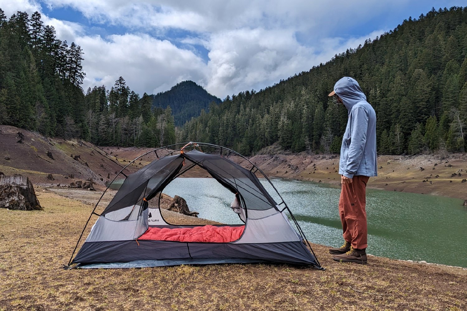 A hiker standing next to the set up REI Half Dome 2 with the rainfly off and the doors open - you can see a red sleeping bag inside and there's a lake a mountain and a pine forest in the background