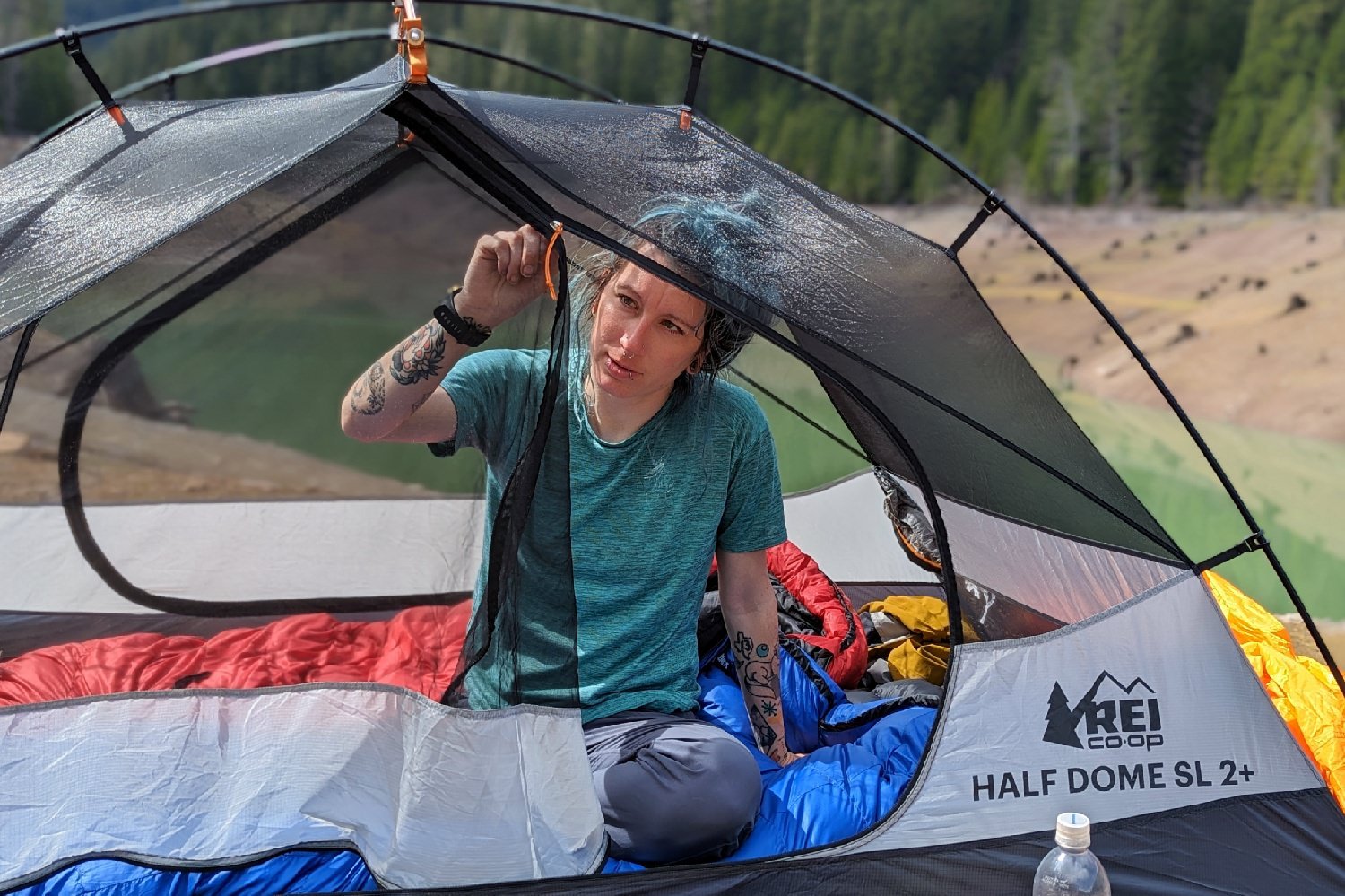 A hiker sitting inside the REI half Dome SL 2 - the door zipper is half open and they are zipping it closed with one hand
