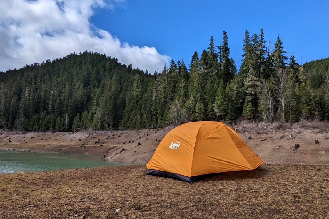 The REI Half Dome SL 2 tent set up in a campsite with a lake and pine forest in the background