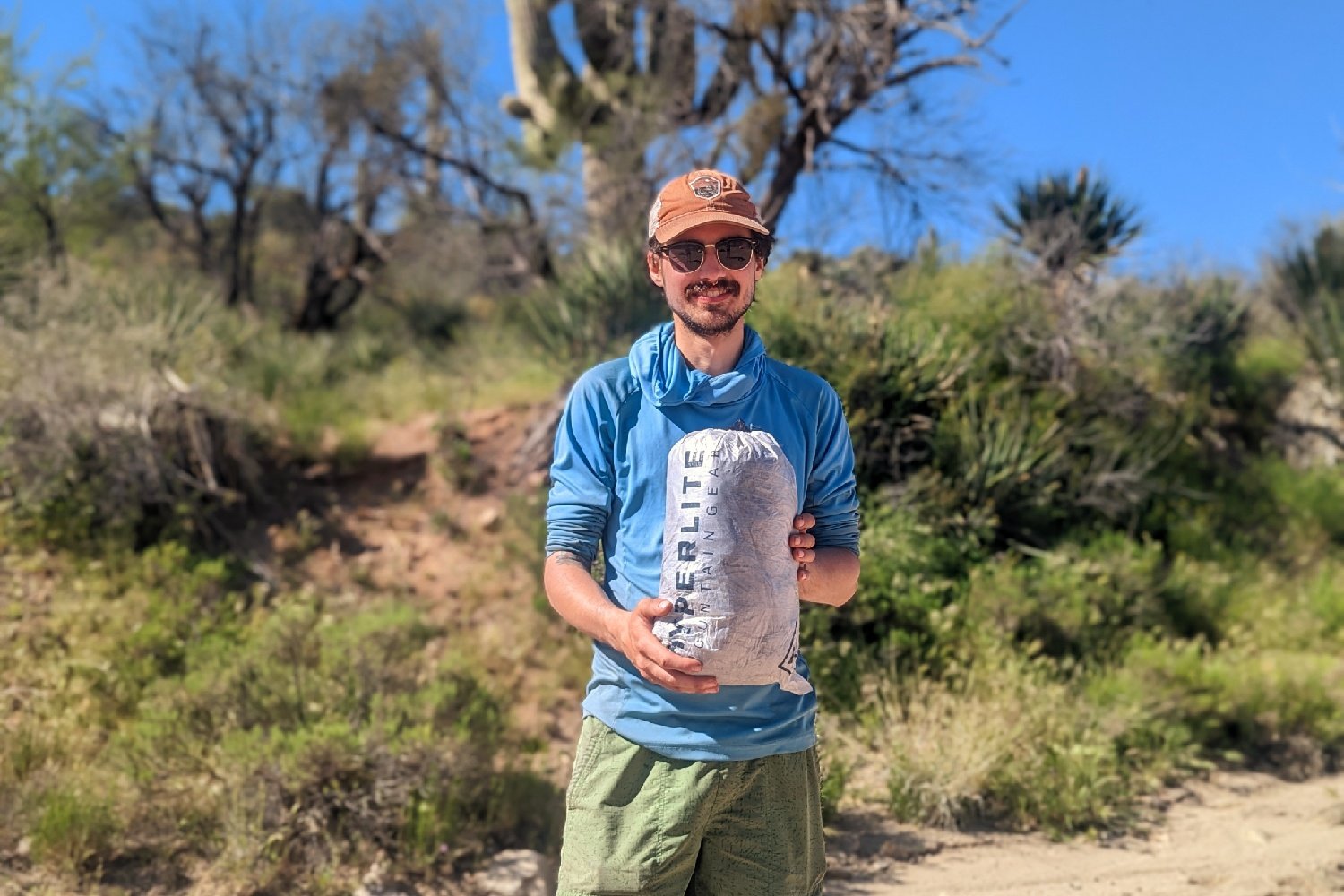 A smiling hiker standing amongst some cacti holding the HMG Unbound 2 tent packed up inside its stuff sack