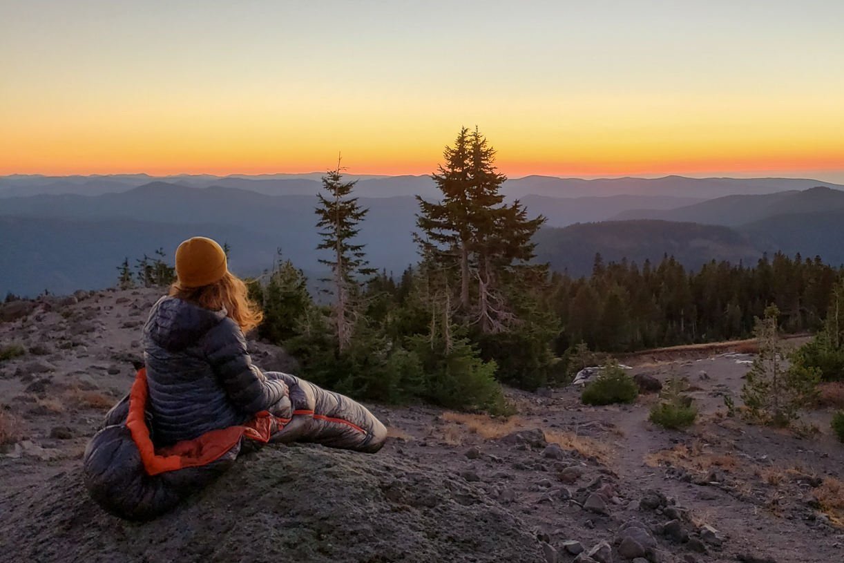 A hiker sitting in an REI Magma 15 sleeping bag looking off at distant sunset and cascading mountain peaks