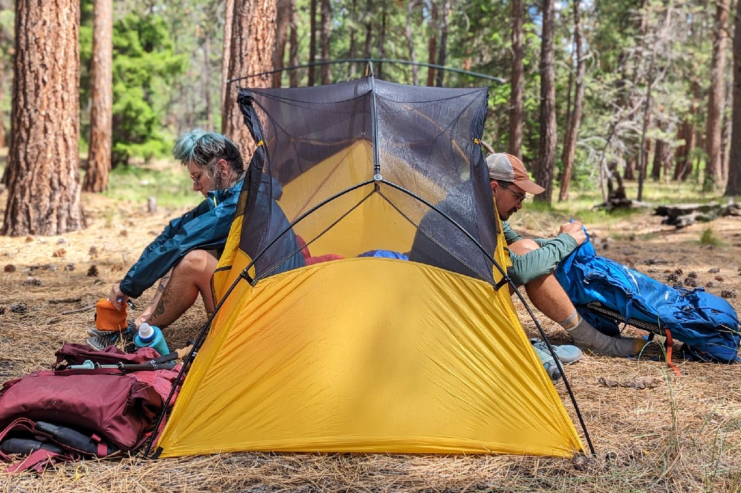 An outside view of the REI Flash 2 Tent from the foot of the tent - two hikers are sitting inside with their legs out either door - one is interacting with a backpack and the other has a cookset in their hands