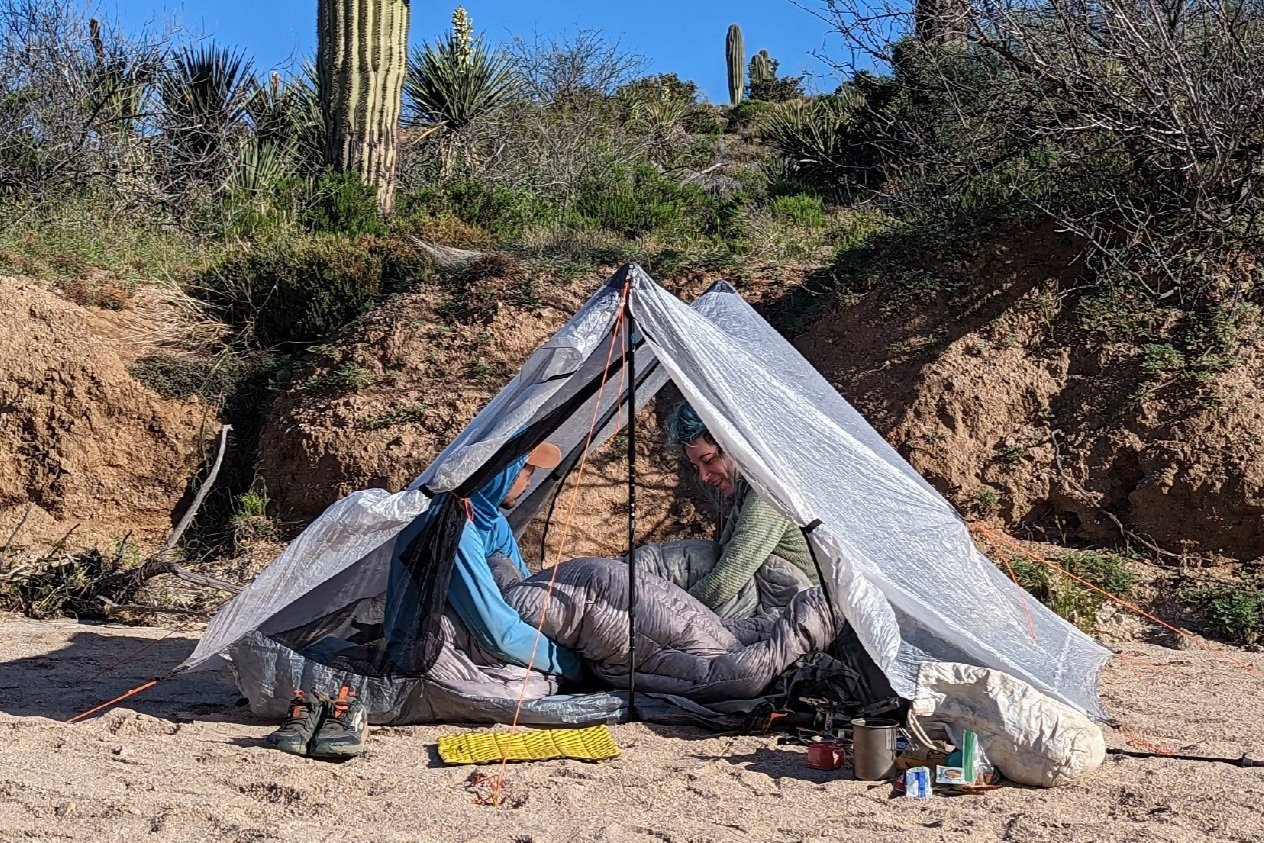 Two hikers sitting in a tent together in a desert campsite with cacti in the background