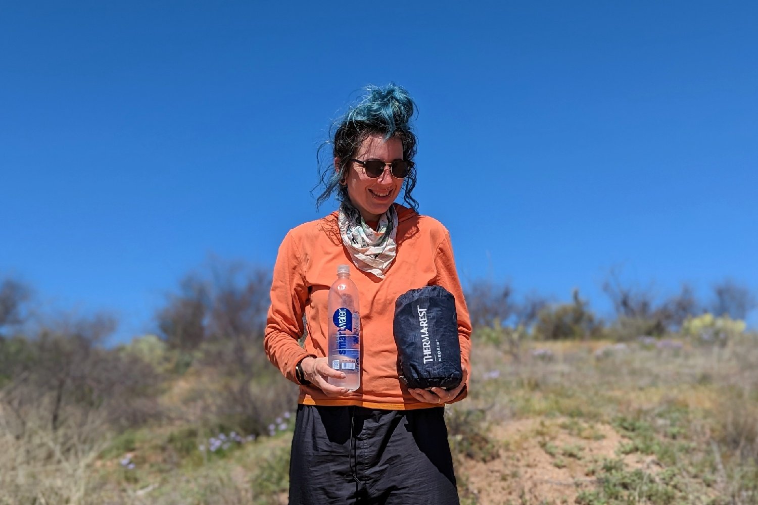 A hiker holding the Therm-a-Rest NeoAir Xtherm NXT packed up in its stuff sack in one hand and a one liter smart water bottle in the other - there's ablurred out desert landscape in the background