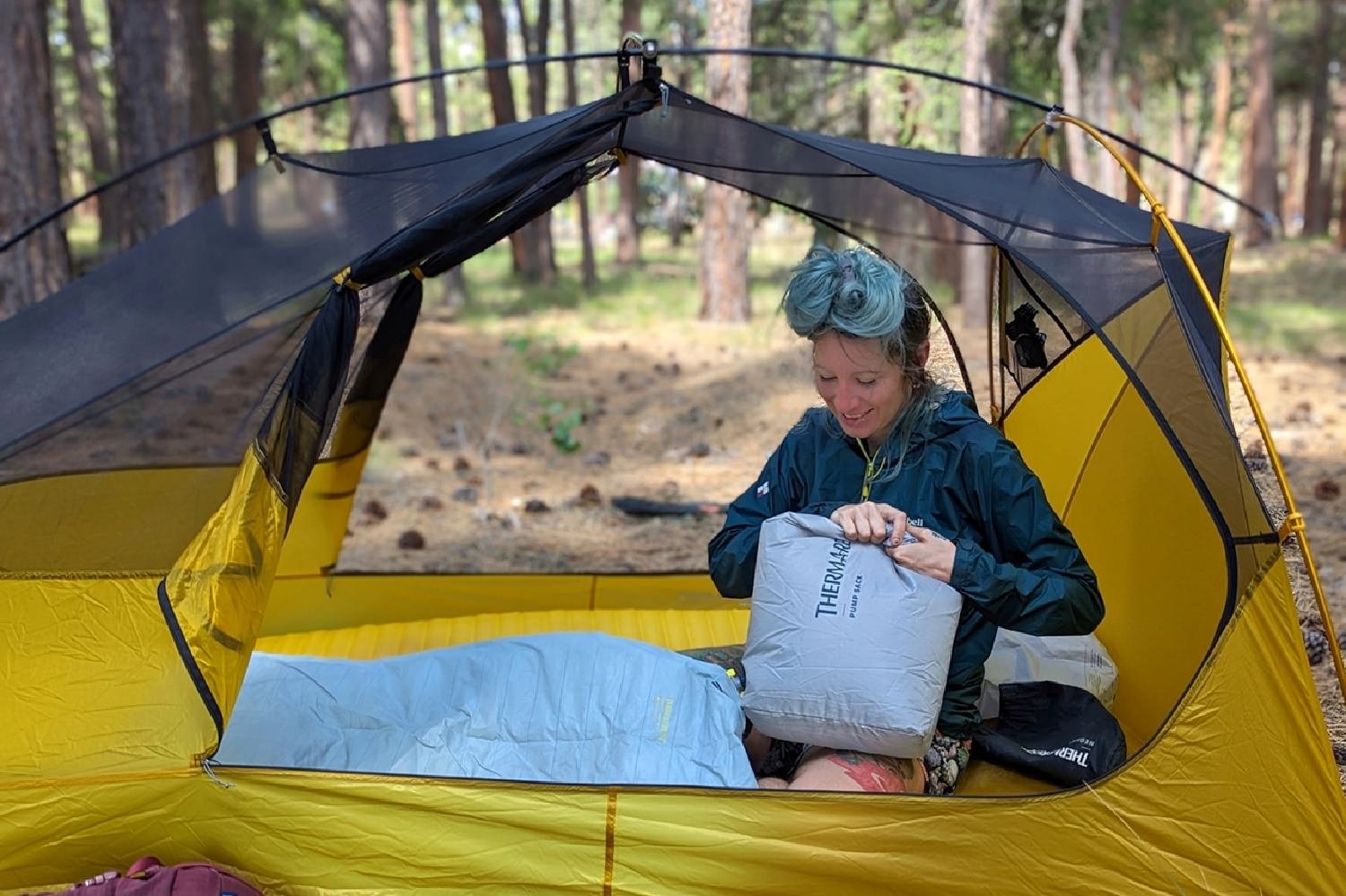 A smiling hiker sitting in a tent in a forested campsite blowing up the Therm-a-Rest NeoAir Xtherm NXT with the included pump sack