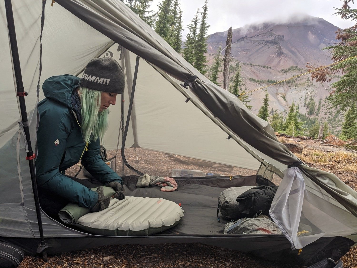 A hiker sitting in a tent and rolling air out fo the Therm-a-Rest NeoAir Xtherm NXT - there is a big mountain in the background and a storm is rolling in
