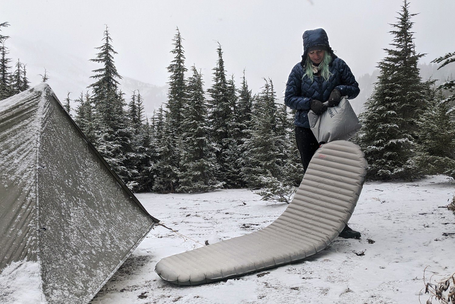 A hiker in blowing up the Therm-a-Rest NeoAir Xtherm NXT using the included pump sack - the hiker is in a very snowy campsite with some pine trees and grey skies