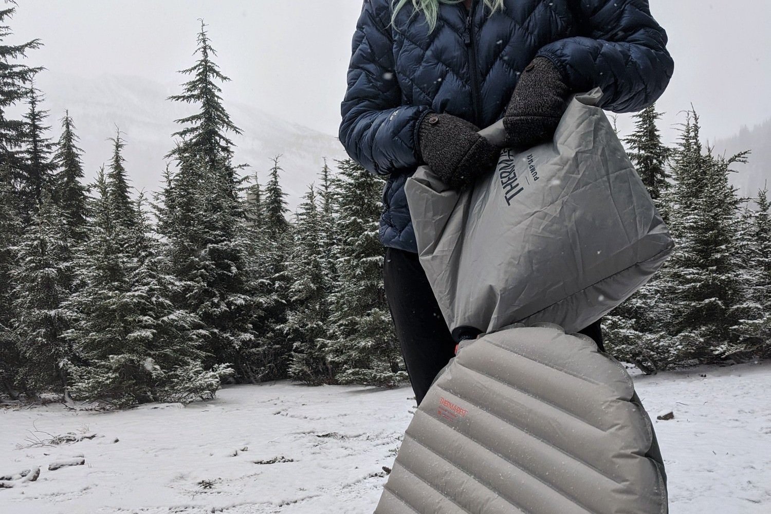 A torso down view of a hiker using a pump sack to inflate the Therm-a-Rest NeoAir Xtherm NXT in a snowy campsite with pine trees