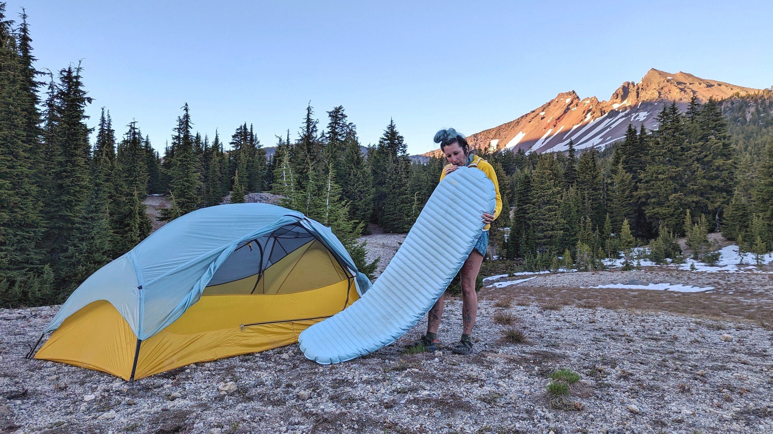A hiker blowing up the Therm-a-Rest NeoAir Xtherm NXT by mouth next to their tent with a mountain in the background