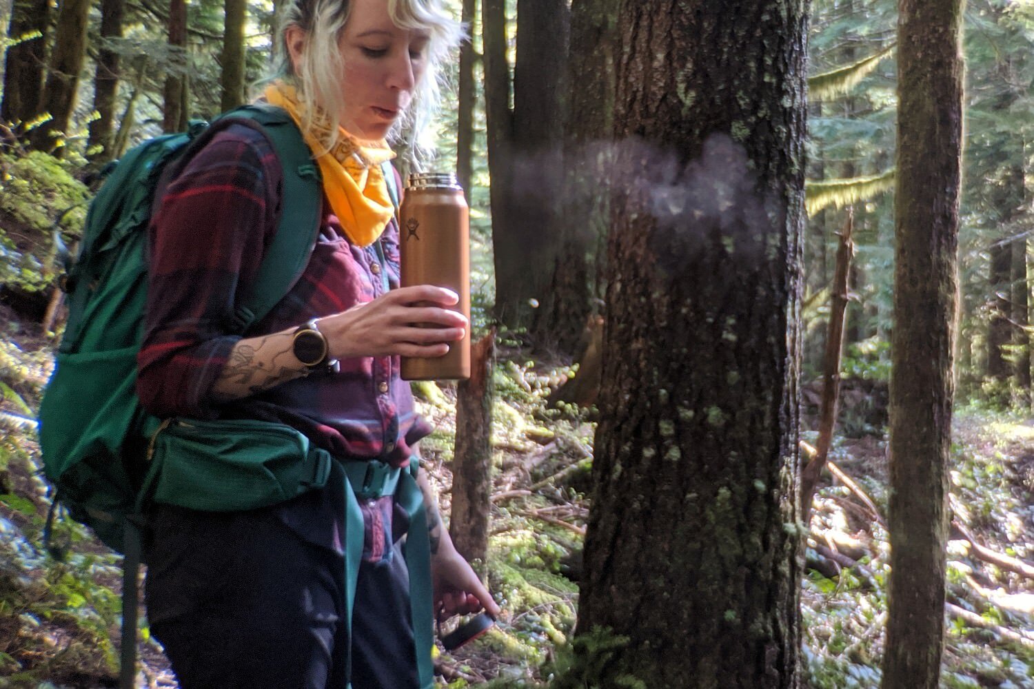 A hiker wearing the REI Trail 40 in a forest - theyre holding a Hydro Flask with a steamy drink in it