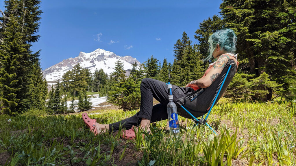 A hiker sitting in a meadow in the Helinox Chair Zero backpacking chair with a snowy mountain peak in the distance