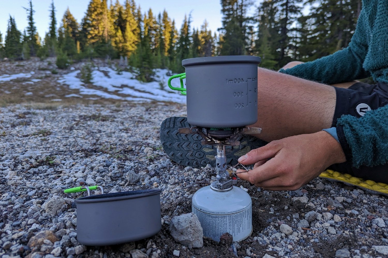 A hiker adjusting the flame on the SOTO Windmaster stove in a rockycampsite with pine trees in the background