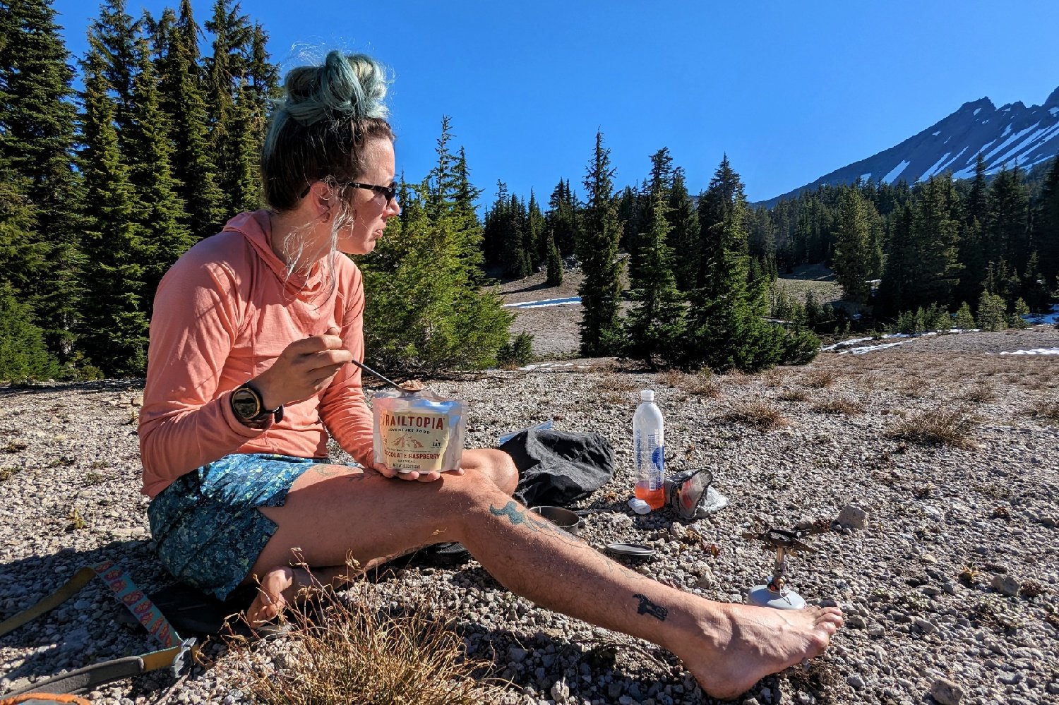 a hiker sitting in a rocky campsite eating food out of a freeze dried meal pouch - the hiker is looking off at a distant peak and the SOTO Windmaster stove is next to them