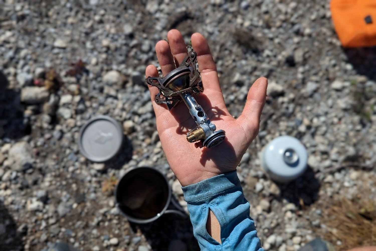 The SOTO Windmaster stove in a hikers hand to show the relative size - the stove is about the size of the hikers palm - there is some backpacking cookgear out of focus in the background on rocky ground