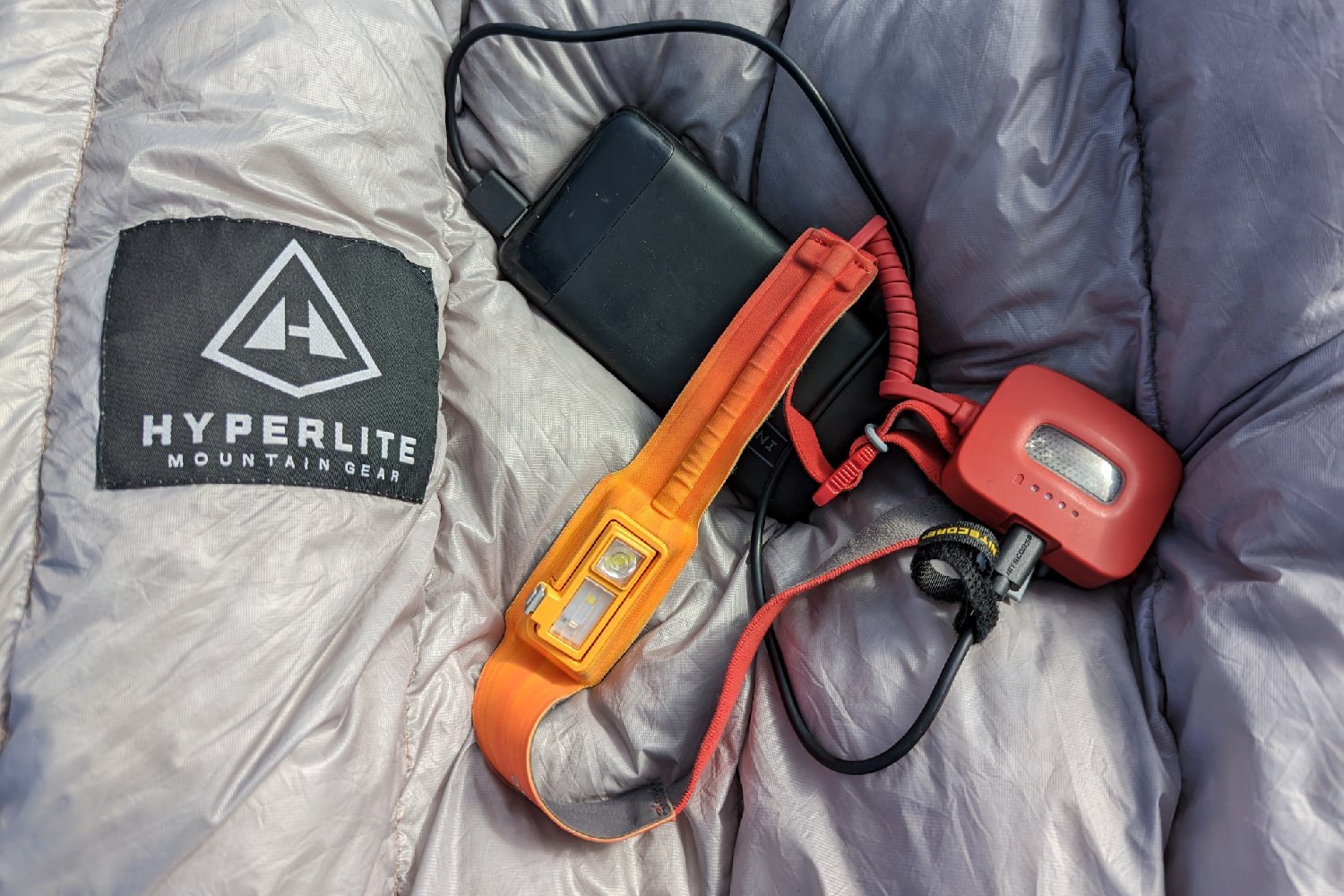 A red Biolite HeadLamp 325 sitting on top of a backpacking quilt. The headlamp is plugged into a INUI 20000 power bank to charge