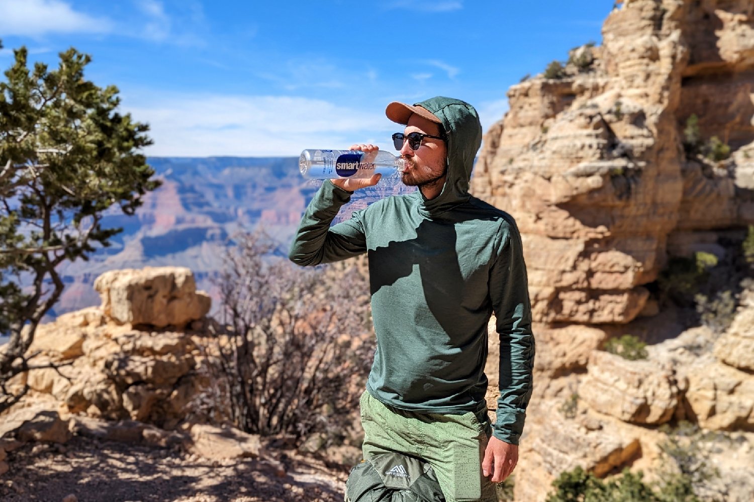 A hiker wearing the REI Sahara Shade Sun Hoodie with the hood up drinking water with a view of the Grand Canyon in the background