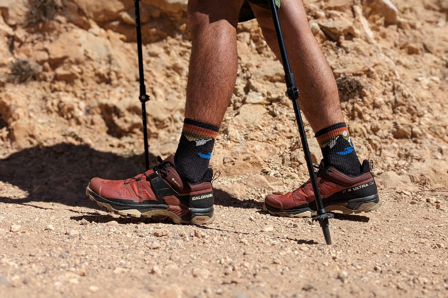 A close up view of a hikers from the knee down wearing Salomon X Ultra 4 hiking shoes on a sandy trail. There's a rocky canyon wall in the background.