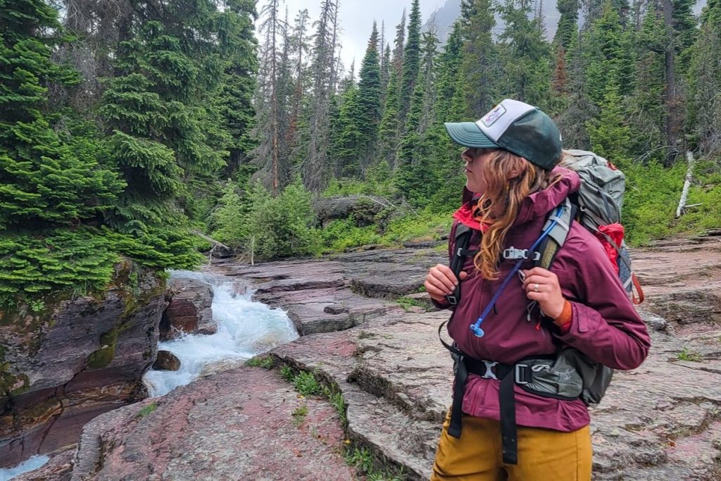 A female backpacker wearing the Rab Kinetic 2.0 rain jacket  near a roaring creek in Glacier National Park
