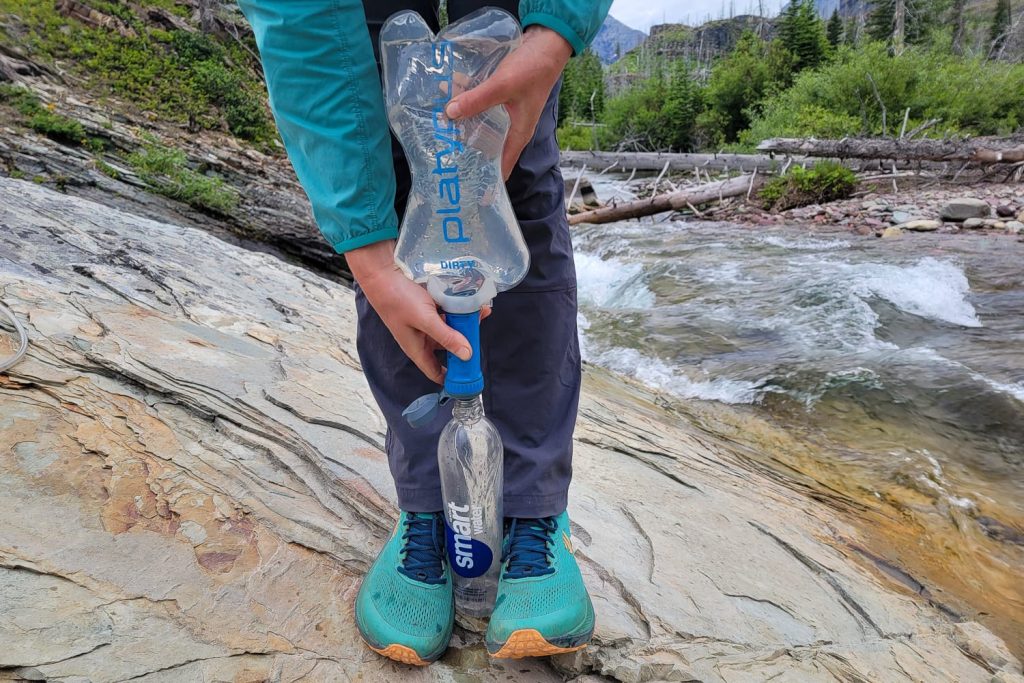 A closeup view of a hiker filtering water with a Platypus Quickdraw Water Filter. There is a fast-moving stream and some pine trees in the background.