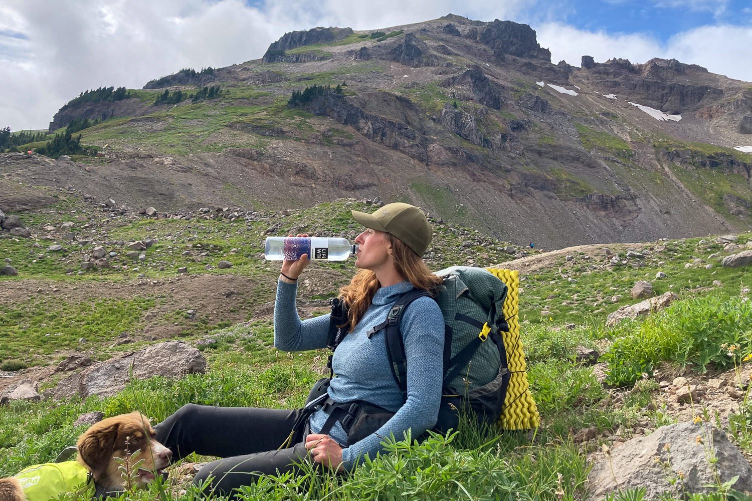 A backpacker sitting for a break on the ground drinking from a water bottle. The hiker is wearing a Patagonia Capilene Air Hoodie and there is a rocky mountain in the background