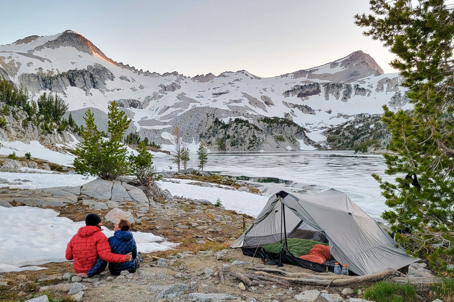 A backpacker couple sitting next to the Lunar Duo tent near a snowy lake at the foot of Eagle Cap Mountain