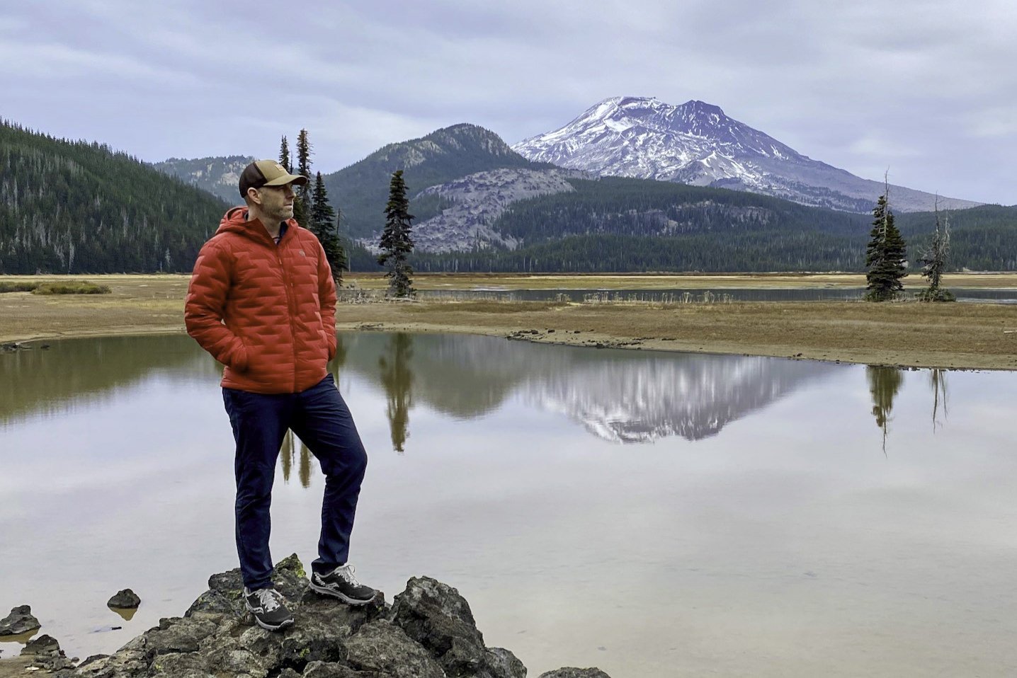 A hiker wearing the Mountain Hardwear Stretch Ozonic Down Jacket in front of a lake at the base of a mountain