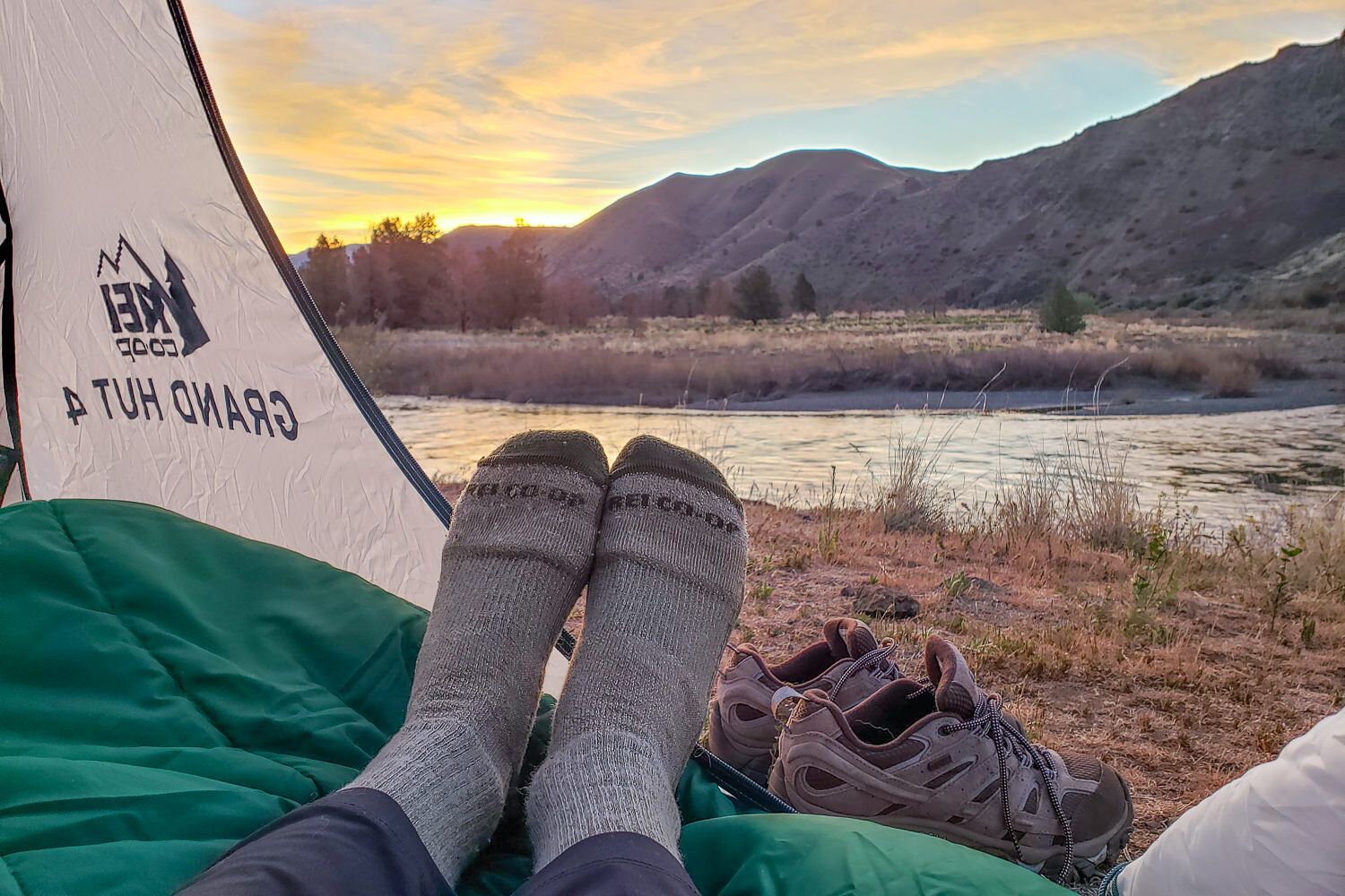 Photo of a person in a tent with their feet in socks. Socks are framed in the open tent door in front of a beautiful lake/mountain sunset