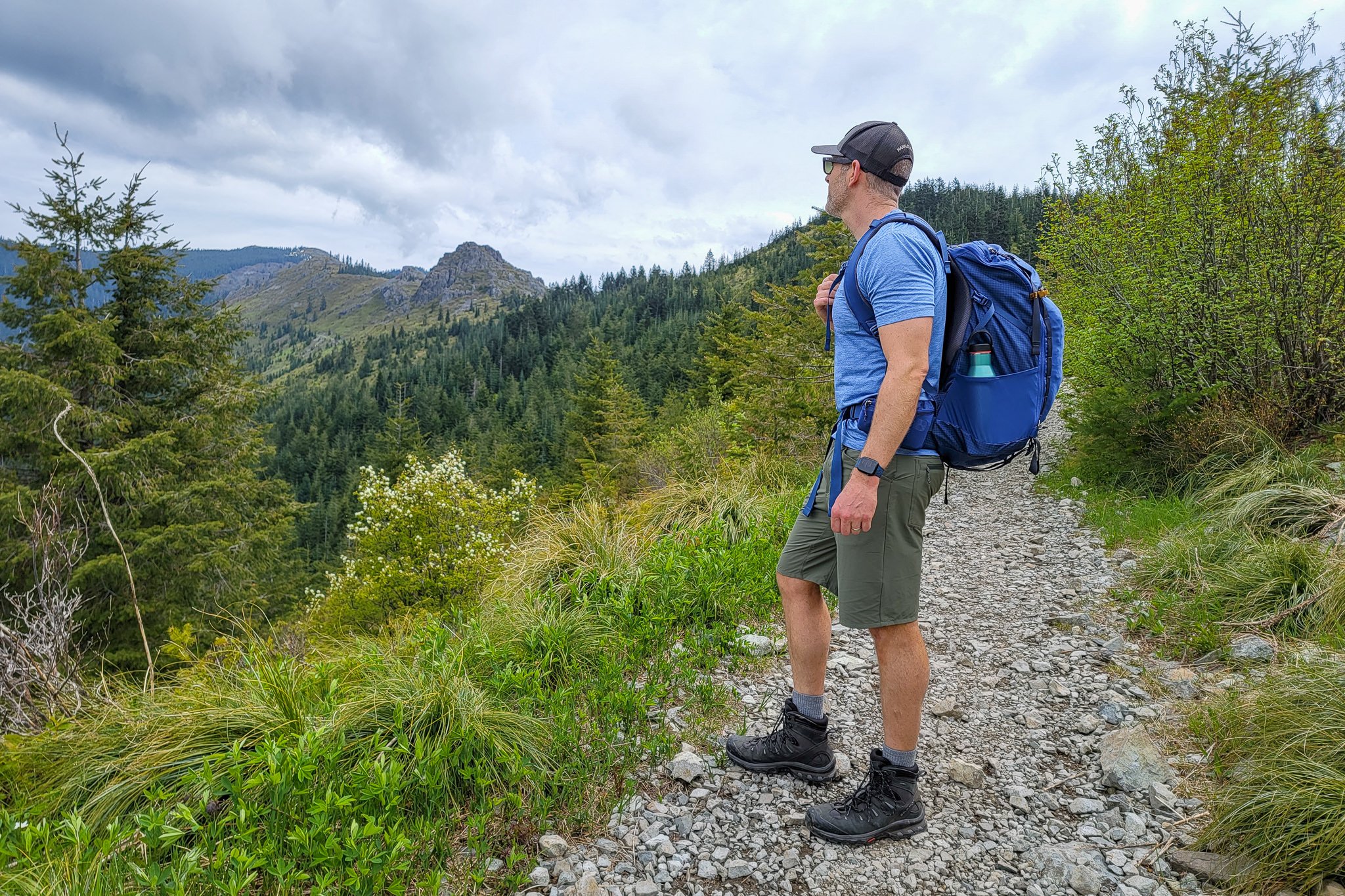 A male hiker standing on a rocky trail wearing the REI Trail 40 backpack looking out at distant mountains under a cloudy sky