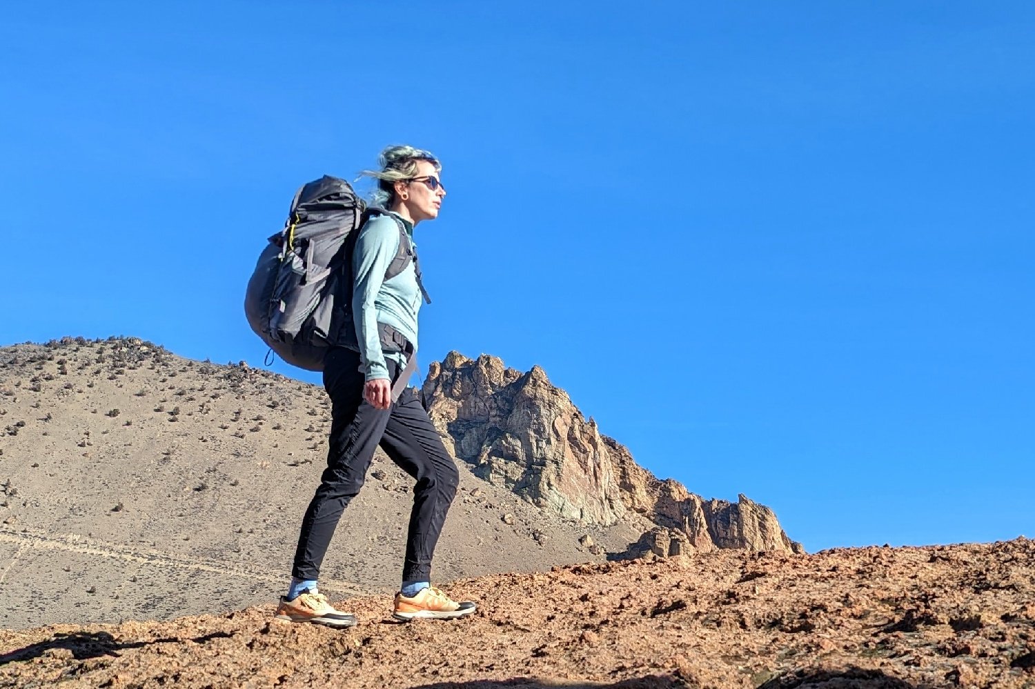 A hiker wearing the REI Flash 55 backpack walking on a ridge trail with a mountain peak and a very blue sky in the background