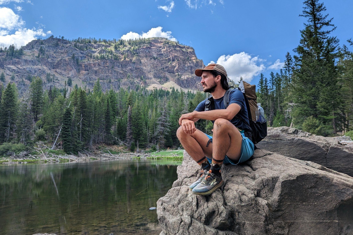 A hiker wearing Altra Lone Peak 7 trail running shoes. He's sitting on a rock in front of a lake looking out into the distance, and there are pine trees and a mountain in the background