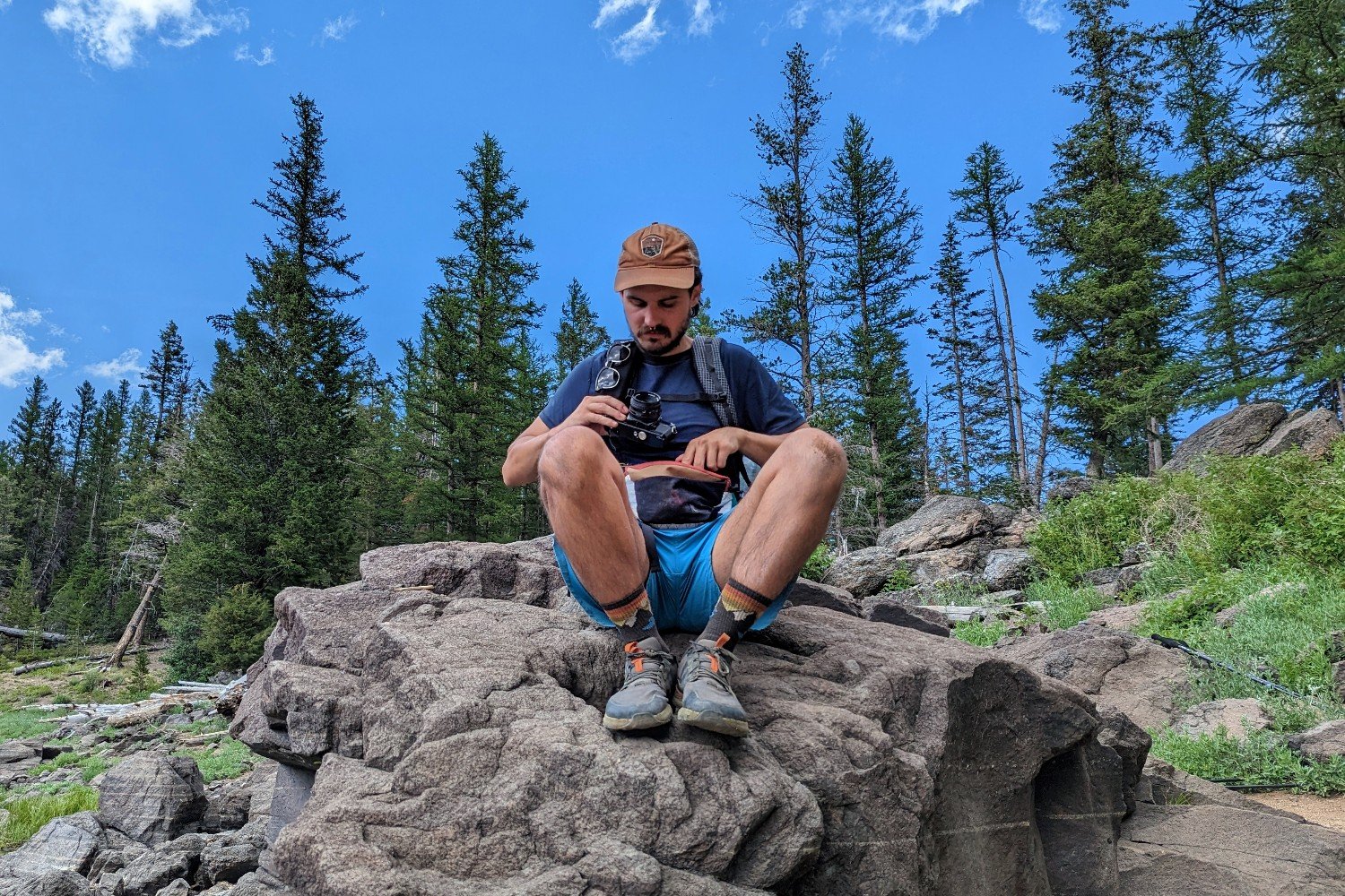 A hiker sitting on a large rock pulling a camera out of an Atom Roo fanny pack. There are pine trees in the background with a bright blue sky and little whispy clouds