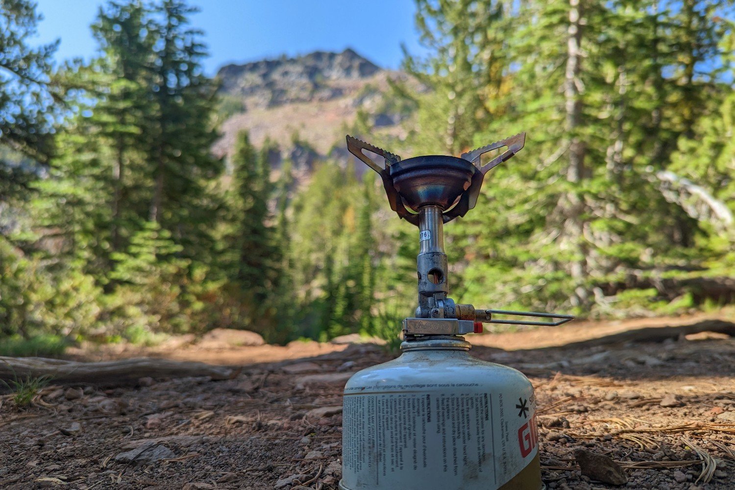 The SOTO Windmaster stove with TriFlex pot support attached to a fuel canister - its a wooded campsite scene with a rocky mountain in the background